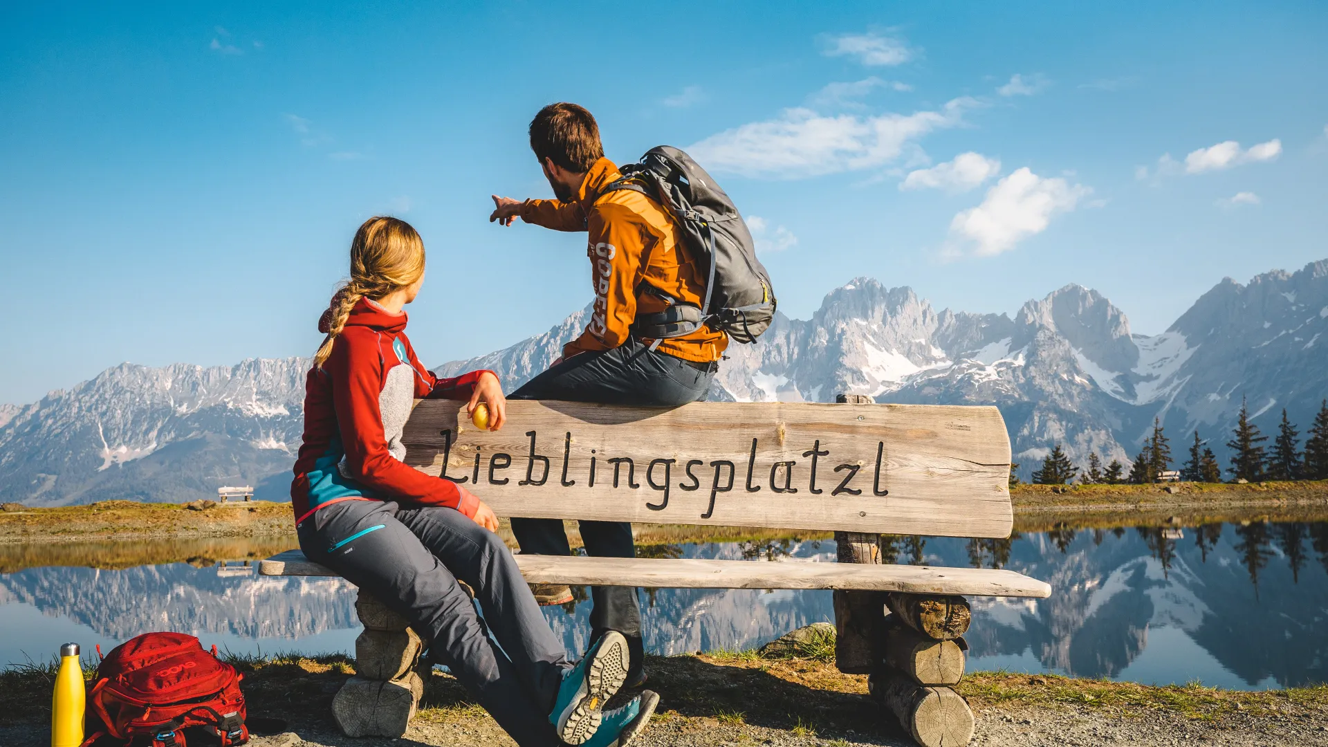 Zwei Wanderer sitzen auf einer Bank mit der Aufschrift 'Lieblingsplatzl' am Astbergsee.