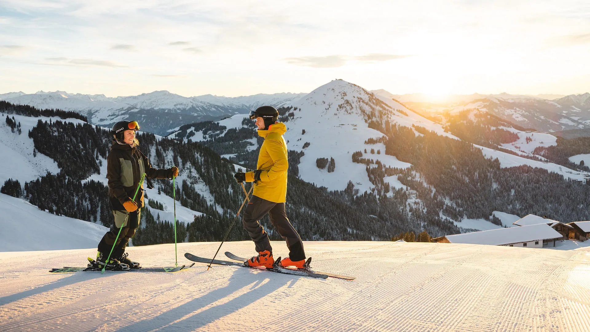 Zwei Skifahrer stehen auf einer Piste in den Bergen bei Sonnenuntergang.